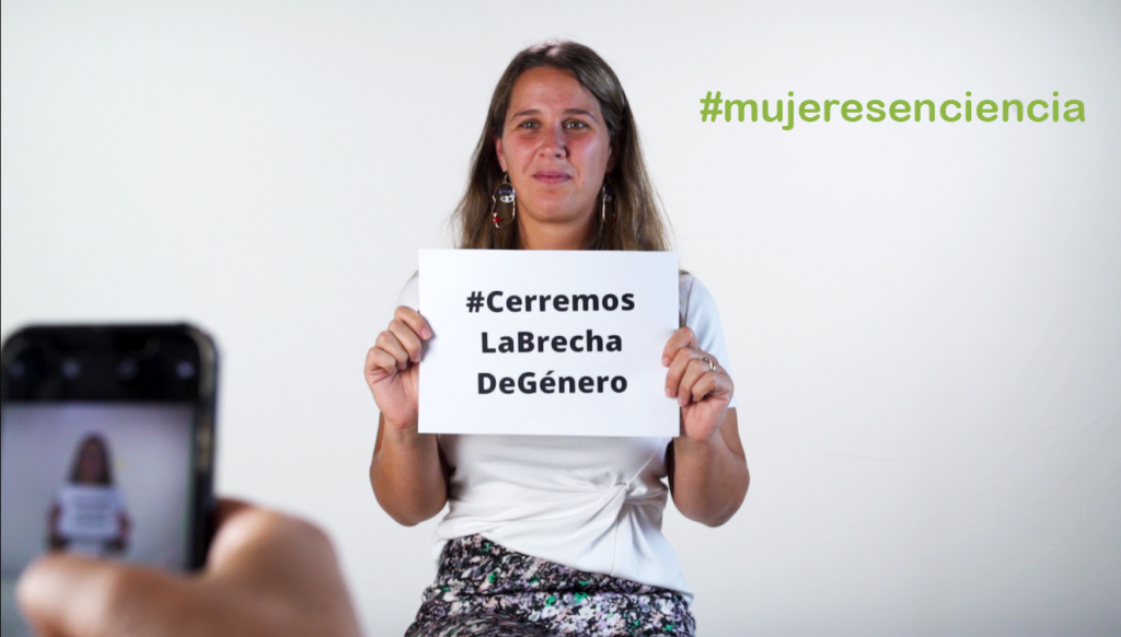Una mujer sostiene un cartel con la etiqueta #CerramoslaBrechadeGenero; al fondo, #MujeresenCiencia